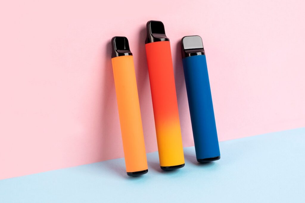 Drei farbenfrohe Einweg-Vapes in Orange, Rot und Blau stehen vor einem pastellfarbenen Hintergrund aus Rosa und Hellblau, modern und minimalistisch inszeniert.